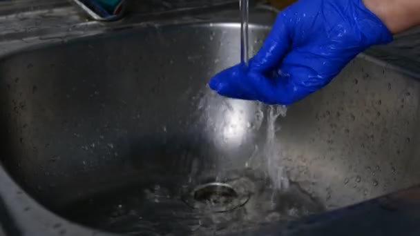 Menschliche Hand in blauem Gummihandschuh wäscht seine Hand unter Wasserstrahl aus dem Wasserhahn in Küche oder Bad. Nahaufnahme. — Stockvideo