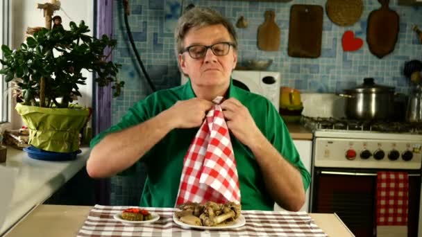Hungrig äldre vuxen man, binder servetten runt halsen, med nöje äter fläskkött vid bordet i sitt hemkök. Medelstor plan. — Stockvideo
