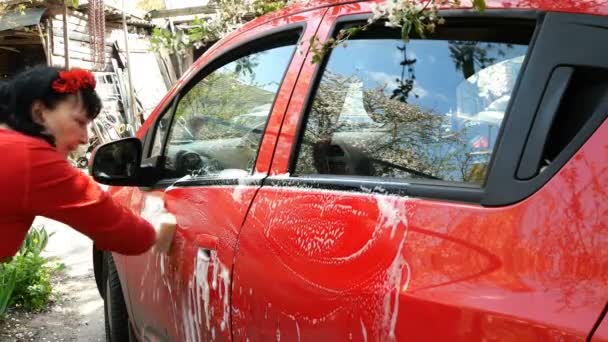 Kırmızı elbiseli bir kadın kirli kırmızı arabayı sünger ve sabunlu köpükle elleriyle yıkıyor. Evinin arka bahçesinde. Ortam planı. — Stok video