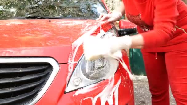 Mujer en ropa roja lava el coche rojo sucio a mano con esponja y espuma jabonosa. En el patio trasero de su casa. De cerca. . — Vídeos de Stock