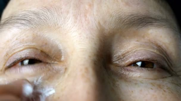Volwassen volwassen vrouw, blanke etniciteit smeren hydraterende cosmetische crème rimpels op haar gezicht met vingers, op zoek naar camera. Close-up. — Stockvideo