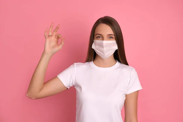 Mutlu esmer kız Corona virüsünden korunmak için beyaz bir maske takıyor, sağlığını ve güvenliğini önemsiyor, güzel bir gülümsemesi var, kendini izole etmeye devam ediyor, olumlu olmak için iyi bir yüzük hareketi gösteriyor. Covid 19