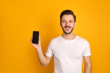 Beyaz tişörtlü mutlu genç adam sarı arka planda izole edilmiş, akıllı telefon sunuyor, kameraya gülümsüyor.