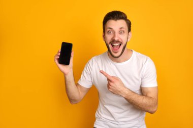 Beyaz tişörtlü komik genç adam sarı arka planda izole edilmiş, akıllı telefon sunuyor, ağzı açık gülüyor, heyecanlanıyor..