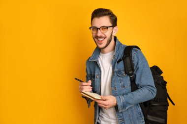 Gözlüklü neşeli bir gazeteci, sarı arka planda beyaz bir tişört ve kot ceket giyiyor, siyah çantasını omzunun üstünde tutuyor, kalemle not defterine yazıyor, gülüyor.