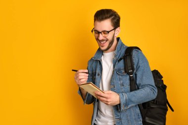 Gözlüklü neşeli bir gazeteci, sarı arka planda beyaz bir tişört ve kot ceket giyiyor, siyah çantasını omzunda tutuyor, kalemle not defterine yazıyor, gülüyor, defterine bakıyor.