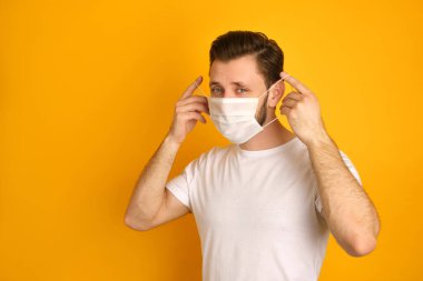 Caucasian young guy puts face white mask on for protecting against covid19, wants to stay safe, has a white t-shirt on, feels calm, holding a mack with his hands clipart