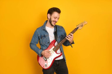 Elektro gitar çalan, gülümseyen, bando çalan, müzik çalan, sarı arka planda, beyaz tişörtlü ve kot ceketli yakışıklı bir adamın portresi.