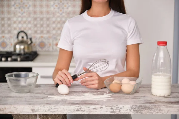 unrecognizable woman cook in white clothes is going to cook in the kitchen, holds a whisk. on the table is a bowl with eggs, a bottle of milk and a flour. wood texture