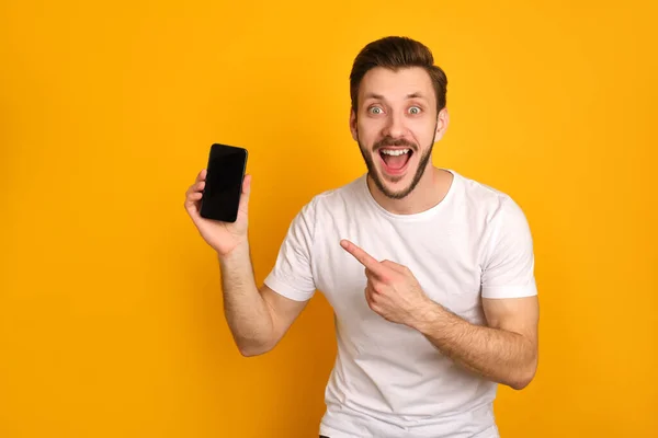 Beyaz tişörtlü komik genç adam sarı arka planda izole edilmiş, akıllı telefon sunuyor, ağzı açık gülüyor, heyecanlanıyor..