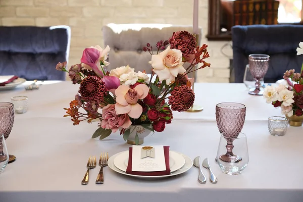 Luxury cozy autumn wedding table decoration in the restaurant. Fresh and dried flowers, roses, carnations. Beautiful table setting: golden appliances, fork and knife, calligraphy guest seating card.