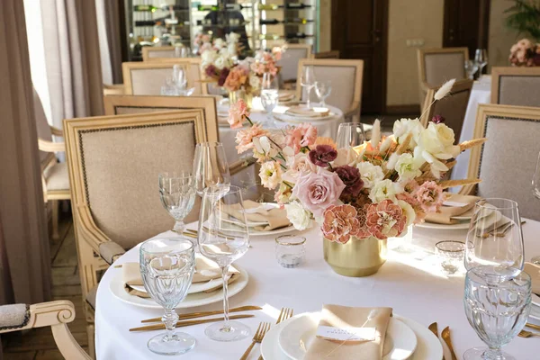 Luxury cozy autumn wedding table decoration in the restaurant. Fresh and dried flowers, roses, carnations. Beautiful table setting: golden appliances, fork and knife, calligraphy guest seating card.