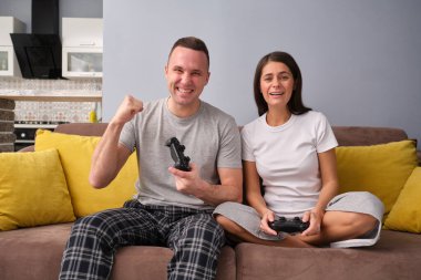 Mutlu genç çift rahat, parlak bir dairede rahat rahat rahat giyiniyor, video oyunları oynuyor, arka planda mutfak, bir adam yumruğunu sıkıyor, eğleniyor, evde vakit geçiriyor.