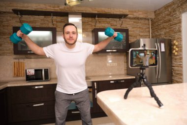 Spor giyim sektöründeki atletik blogcu, mutfakta egzersiz yaparken telefonda video çekiyor. Ev aktivitelerinde kal. Pandemiden enfeksiyonu önlemek için..