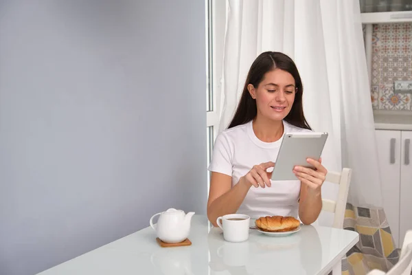 Uzun saçlı esmer kız mutfakta kruvasanla çay içiyor, beyaz tişört giyiyor, arka planda beyaz perdeler var, tablette e-kitap okuyor, kendini izole ederek iyi vakit geçiriyor.