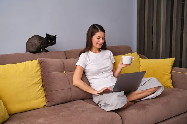 Kahverengi kanepede oturan esmer bir kadın, oturma odasında sarı yastıklı, pijama giyen, elinde çay tutan, dizüstü bilgisayarında komik videolar izleyen, eğlenen, siyah bir kedi kameraya bakıyor.