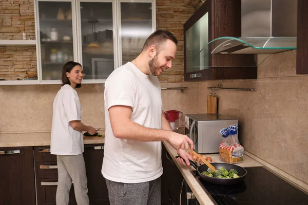 Mutlu genç çift kahvaltı hazırlıyor, adam brokoli kızartıyor, çırpılmış yumurta pişiriyor. Kız taze sebzeleri dilimliyor. Koronovirüs pandemik covid-19 sırasındaki kendini izolasyon..