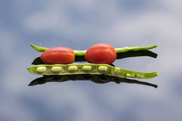 Green beans with their seeds and red cherry tomatoes on a glass surface that reflects the sky.