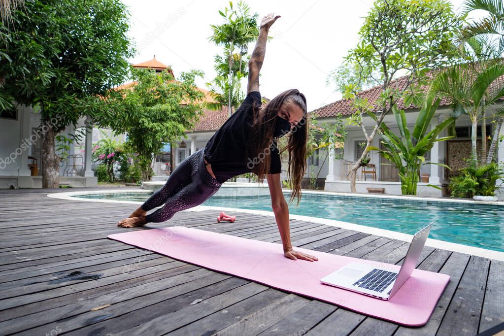 young woman in protective mask practicing yoga near swimming pool