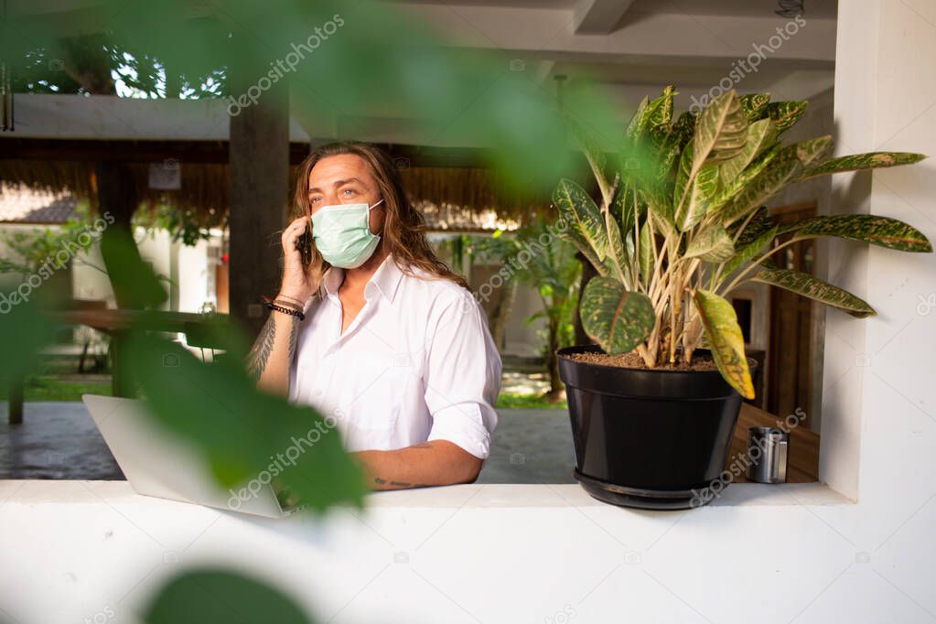 handsome young man in medical face mask working at home with laptop on summer terrace background