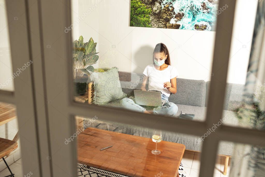 young woman working at home in face mask in room sofa background