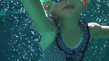 Çizgili mayo ve gözlüklü küçük kız havuzda suyun altında yüzüyor.