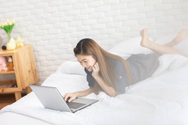 Güzel, çekici, Asyalı genç bir kadın yatağa uzanıyor ve bilgisayarını sosyal medya ve online alışveriş için kullanıyor tatillerde rahat yatak odasında dinleniyor, yatak odasında yaşam tarzı kavramını kullanıyor.