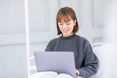 Güzel, çekici, Asyalı genç bir kadın yatağa uzanıyor ve bilgisayarını sosyal medya ve online alışveriş için kullanıyor. Tatilde yatak odasında rahatlıyor, yatak odasında yaşam tarzı kavramını kullanıyor.