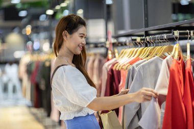 Güzel, çekici, Asyalı genç bir kadın elbise mağazasında rafta asılı duruyor. Mutluluktan uçuyor ve alışveriş merkezinde, seçici odak noktasında, moda ve alışveriş konseptinde eğleniyor.