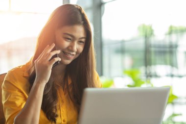 Dizüstü bilgisayarlı gülümseyen ve akıllı telefondan müşteriyle konuşan güzel Asyalı kadın kahve dükkanının dışında, Business Startup Concept 'da başarılı bir iş için fikir ve gereksinim edinmek için çalışıyor.