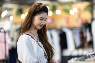 Güzel, çekici, Asyalı genç bir kadın elbise mağazasında rafta asılı duruyor. Mutluluktan uçuyor ve alışveriş merkezinde, seçici odak noktasında, moda ve alışveriş konseptinde eğleniyor.