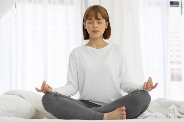 Çekici Asyalı kadın yoga nilüferine poz veriyor. Sabah uyandıktan sonra yatakta meditasyon yapıyor. Kendini çok rahat ve rahat hissediyor.