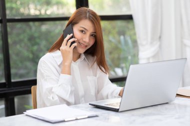 Dizüstü bilgisayarlı gülümseyen ve akıllı telefondan müşteriyle konuşan güzel Asyalı kadın kahve dükkanının önünde, Business Concept 'da başarılı bir iş için fikir ve gereksinim edinmek için çalışıyor.