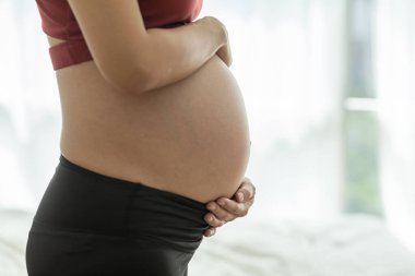 Mutlu, Güzel, Çekici, hamile, Asyalı kadın. Yatakta oturuyor, gülümsüyor ve karnını tutuyor. Çok gururlu ve sevgi dolu.