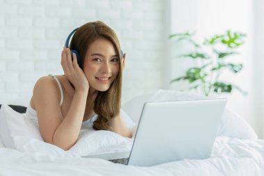 Güzel, çekici, Asyalı genç bir kadın müzik dinler ve yatakta yatar. Yatak odasında dizüstü bilgisayara bakar. Kendini rahat ve rahat hisseder.