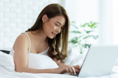Güzel, çekici, Asyalı genç bir kadın yatağa uzanıyor ve bilgisayarını sosyal medya ve online alışveriş için kullanıyor tatilde rahat bir yatak odasında dinleniyor, evde yaşam tarzı kavramını kullanıyor. 
