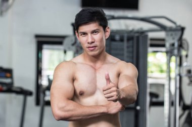 Güçlü Atletik Asyalı adam kollarını kavuşturmuş, spor salonunda kameraya bakıyor. Sağlıklı olmak için başparmağını kaldırıyor. Kaslı adam kendini güçlü ve sağlıklı hissediyor.