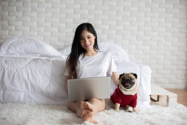 Dizüstü bilgisayarda köpek Pug cinsi köpeklerle yazan ve çalışan bir kadın dizlerinin üzerine çöküp ekrana bakıyor ve mutluluk ve dostça bir konsept hissediyor.