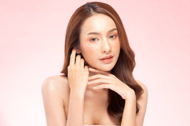 Güzel Asyalı kadın, yumuşak yanaklı gülümsemeye, temiz ve taze tenli mutluluğa, olumlu duygulara, pembe arka plana, güzellik kozmetiğine ve kaplıcaya dokunuyor.