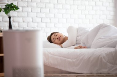 Kadın, temiz hava ve sağlıklı yaşam için, evdeki toz PM2.5 HEPA 'yı filtrelemek ve temizlemek için, rahat beyaz yatak odasında hava temizleyici ile uyur. Hava kirliliği kavramı