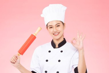 Güzel Asyalı şef kadın gülümsüyor ve pembe arka planda izole edilmiş oklavaları tutuyor, Mutluluk ve Neşeli Profesyonel Şef Konsepti