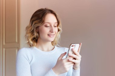 Sarışın kız cep telefonuna gülümsüyor gibi görünüyor. Evde bir odada genç bir kız telefonuna bakıyor..