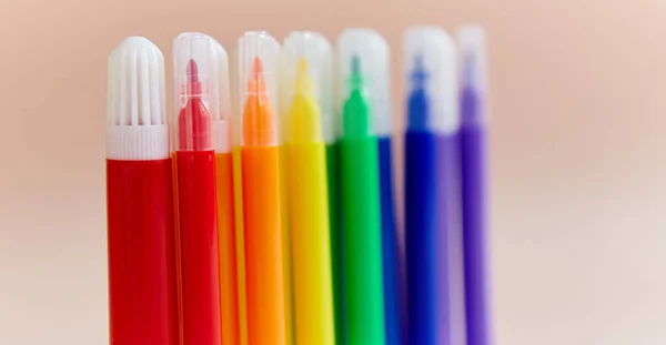 Colored markers thin thick red yellow green pink blue purple light background. Office supplies the color of the rainbow are on the table close up