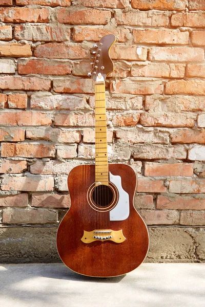An old wooden guitar stands against a brick wall. The sun\'s rays fall on the guitar strings