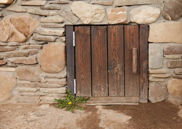 An old wooden door in the stone wall. The door is square with vertical boards and wooden handle wall made of river stone.