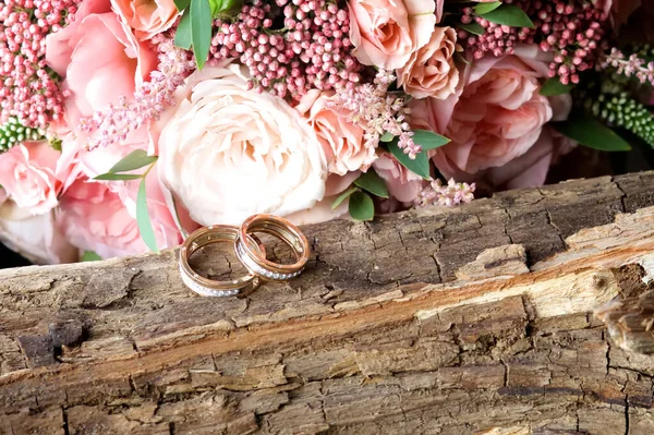 Gelinin buketi pembe çiçeklerin masasında. Buketin yanındaki ağacın kabuğunda iki altın yüzük var..