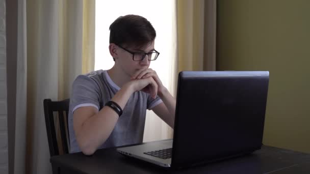 眼鏡をかけた勤勉な10代の若者がテーブルに座ってノートパソコンで宿題をしてインターネット上で情報を勉強しています — ストック動画