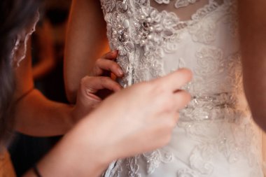 Gelinlik giymiş gelin sırtında duruyor. Kız arkadaşı danteli elbiseye bağlıyor. Elleri kapalı.