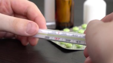 4 bin. Genç adam termometreye bakıyor. Karanlık bir masada haplar ve beyaz kutular var..