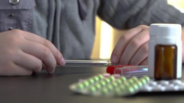 体温計を見てる若者 暗いテーブルの上に丸薬と医療用の白い箱 — ストック動画
